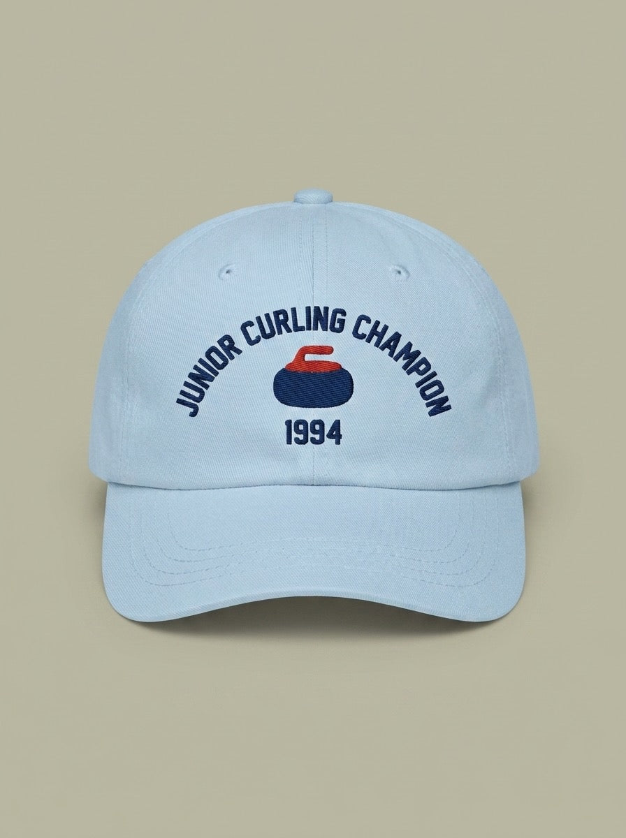 Pale blue dad hat embroidered in navy with JUNIOR CURLING CHAMPION curved above a curling rock, and 1994 below.